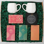 Two white mugs and hot cocoa packets in a decorative box on a light gray background